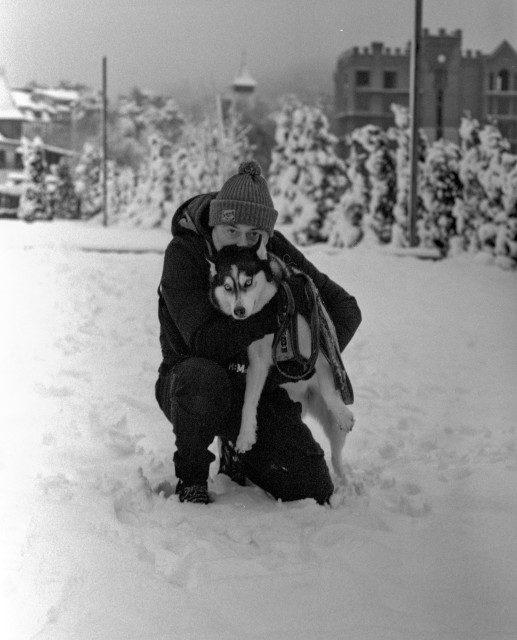 It’s a black-and-white winter portrait showing a person crouching in deep snow while hugging a Husky. The person is bundled up in winter clothes, including a beanie, and leans their face close to the dog’s head. The Husky looks directly at the camera with bright, striking eyes. Behind them, snow-covered trees and blurred buildings create a soft, atmospheric backdrop, giving the scene a calm, quiet, wintry feel.
