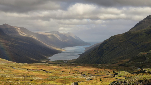 A colour photo of a landscape of steep mountains on either side of a long fjord. Heavy cloud above is creating areas of sunlit rock between the shadows. The foreground is a rocky plateau with golden brown vegetation and streams running through it, brightly lit by sunlight from behind the viewpoint.