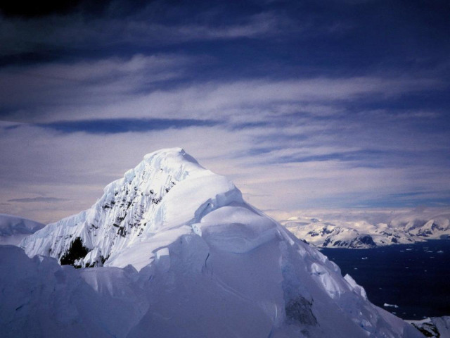 Ein Berggipfel mit viel Schnee uns Eis im Vordergrund. rechts dahinter ist etwas Meer zu sehen und weiter dahinter flachere Berge in Schnee und Eis gehüllt. Blauer Himmel mit dünneren weißen Wolken.