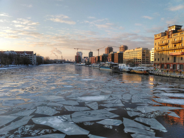 Farbfoto im Querformat. Zu sehen ist der Blick von der Oberbaumbrücke Richtung Berlin-Mitte. Das Licht ist durch den Sonnenuntergang getönt, die Häuser auf der rechten Flussseite leuchten. Auf der Spree treiben viele Eisschollen.