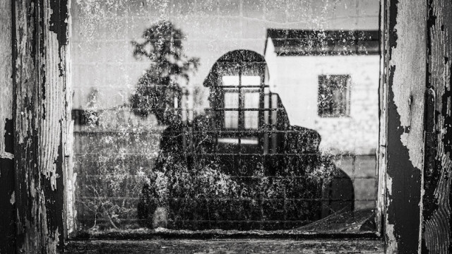 Jemand von außen fotografiert durch eine Scheibe von einem alten Gebäude. Man sieht teilweise den Rahmen vom Fenster. Die Person  spiegelt sich in der alten schmutzigen Scheibe. In dem Bereich wo der Kopf ist, ist ein vergittertes Fenster zu sehen. Dieses stammt von der gegenüberliegenden Seite vom Raum. Das Foto ist in Schwarzweiß.