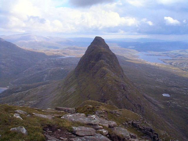 The view east from the top of Suilven, inland from Lochinver on the west coast of Sutherland. The image shows descending rocky ground in the foreground leading to a narrow neck and then climbing to a sharply pointing peak, which is slightly lower than us. In the background is much lower lying land of stony ground with rivers and lochs. The sunlight is patchy.