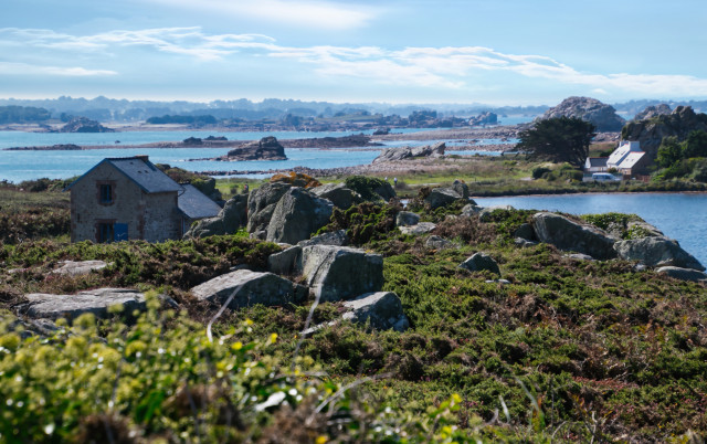 Eine malerische weil schärenreiche Küstenlandschaft mit felsigen Ufern, verstreuter Vegetation und zwei kleinen traditionellen Häusern in der Nähe des Wassers. Die Szene zeigt ein ruhiges blaues Meer und ferne Inseln unter einem teilweise bewölkten Himmel.