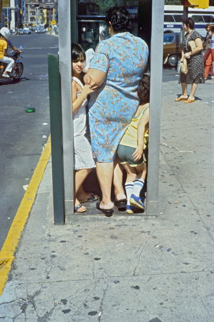 The photo captures a candid moment of a woman and two children squeezed into a public phone booth on a New York City street. 