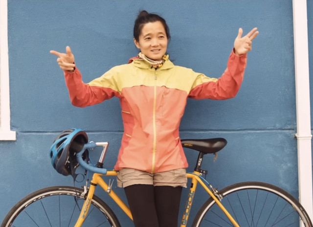 A woman leaning against her bike that’s against a blue outside wall. She’s smiling and gesturing with both hands at the city/world around her.