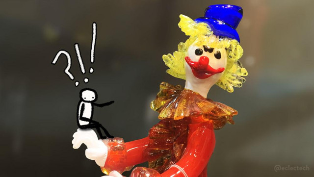 A photo of a glass clown against a blurred background. It has a white face and hands, bright red jacket with orange-red ruff, yellow curly hair and a bright blue hat (we are talking serious primary colours here). It looks to camera with black eyes, frowning brows, red nose and grinning red mouth. On its hand sits a small scribbled figure, also looking to camera but gesturing towards the clown. Above their head is a large question mark and exclamation points.