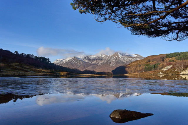 Snow capped peaks of Beinn Eighe under a blue sky reflected in Loch Coulin. Branches of a pine tree intrude into the sky and a rock in the foreground gives a little close interest. 