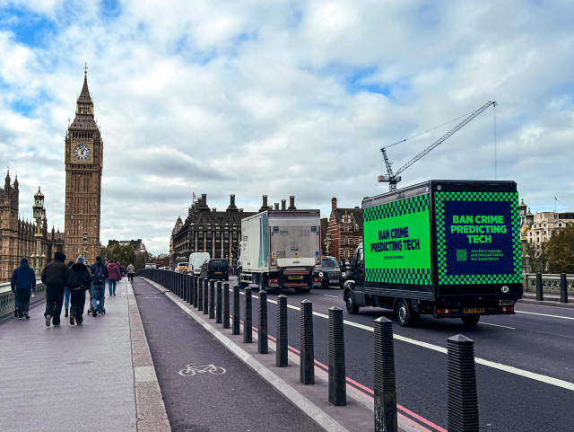 Photo of a van with digital displays reading 'Ban Crime Predicting Tech' on a green background with checkered borders. The van is photographed crossing Westminster Bridge and moving towards Big Ben and the Houses of Parliament in London, UK.