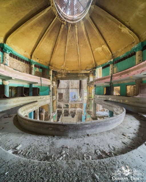 The image shows the decaying, multi-level, horseshoe-shaped auditorium with a partially visible domed ceiling. The structure, once a thriving cultural venue, features faded yellow and teal paint on the walls and balconies