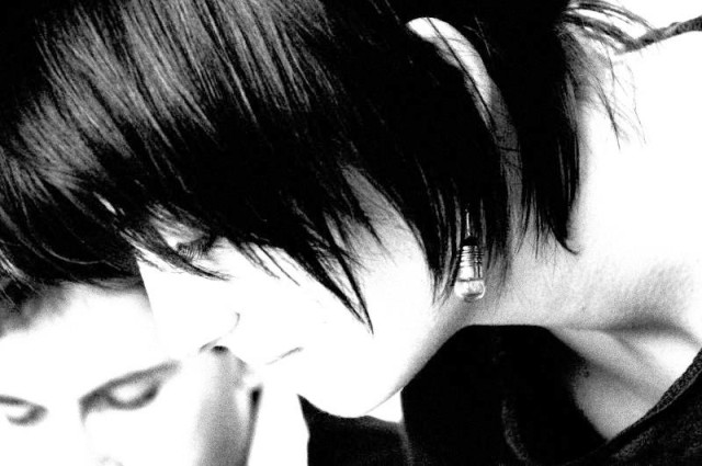 Une photo en noir et blanc, très contrastée, au format paysage . Au premier plan le visage d'une jeune femme de profil, penchée en avant  . Sa chevelure cache en partie ses traits également estompés par le contraste. En arrière plan, à gauche, le haut du visage d'un jeune garçon dont on ne voit qu'un peu de cheveux, le front et la ligne des yeux.  Leurs deux regards ,  captivés, sont tournes vers un point hors champ.