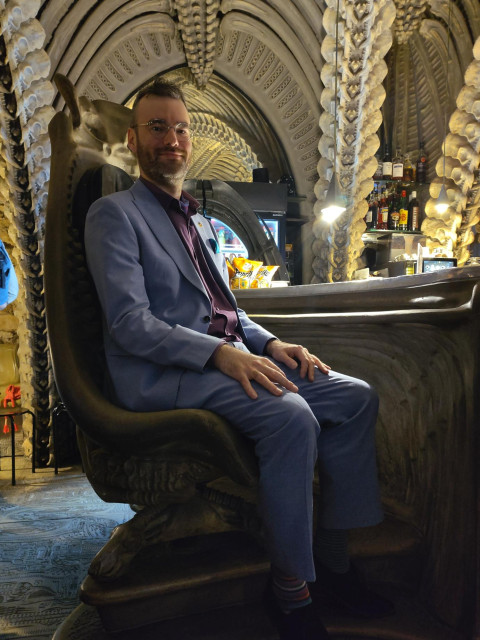 Me, a white man in a suit, sitting in an HR Giger "Harkonnen Chair" designed for the (never made) Jodorowsky 'Dune' movie. The photo was shot at the Giger Bar at the HR Giger Museum in Gruyères, Switzerland.