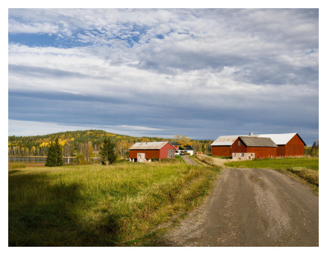 En liggande bild på en gård vid slutet på en grusväg. Gräsfält på båda sidan vägen och gården består ett par boningshus och några ekonomibyggnader. I bakgrunden syns en sjö till vänster och bortom den skog i höstfärger. Himmelen molnig med lite blått.