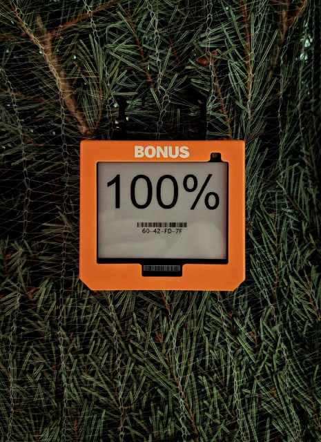 A photo of an electronic shelf label that says “BONUS” at the top and displays “100%” on the screen. It’s hanging in front of a Christmas tree wrapped in netting.