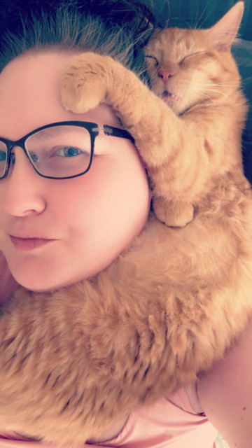 Selfie of me with my orange boy cat, Roux, wrapped around my neck and head sleeping with his mouth open and paws curled on my forehead and his stomach