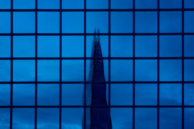 Photo of the top of the Shard, a tall spike of glass and steel building in London, reflected in a grid of square windows. Because the windows aren't perfectly flat, the skyscraper appears to wobble as it passes through four-and-a-bit squares. Because it's blue hour, the Shard and the cloudy sky behind it are blue.