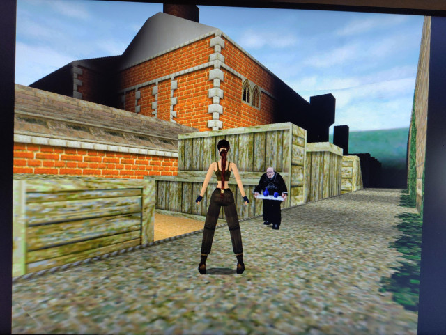 Photo of my desktop computer screen showing Lara Croft and Wilson on the training grounds of Manor Croft. Lara is in a training outfit, Wilson is dressed in black and holds a tray with some tea, the manor is made of orange brick with wooden roofs.