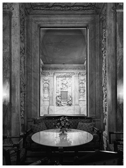 Schwarz-Weiß-Foto im Hochformat mit weißem Rand. Blick aus einem Zimmer im königlichen Palast von Amsterdam. Im Vordergrund ein runder Tisch, dahinter ein Fenster, das auf einen anderen Raum hinausgeht. Er ist mit vielen Reliefs und Statuen aus weißem Marmor dekoriert. 