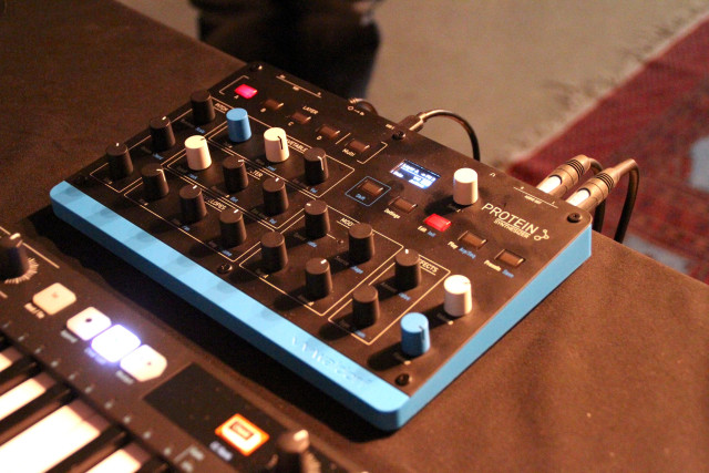 Waldorf Protein tabletop synthesizer with light blue plastic housing and black metal front plate, filled with some 30 colored knobs and buttons.
