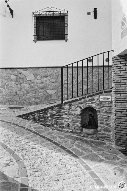 A winding stone pathway leads up to a small fuente inset into a textured stone wall with a black iron railing. Above, a decorative window with burglar bars is set against a white wall, while a small fountain can be seen near the base of the slope.