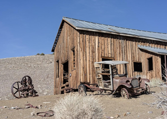 A color landscape photo of a large, very weathered, wood building on a nearly barren hillside. The building was a machine shop. The windows and doors are long gone. The front of the building faces toward the left. Outside, n front of the building, is a large rusted machine with two large gears that look like spokes in a wheel. On the right is a very weathered early pickup truck. It has a rusted chassis, hood , and fenders. The cab of the pickup is wood. Like the building behind the truck, its windows and doors are long gone. The sky is clear and very blue in the high elevation. 