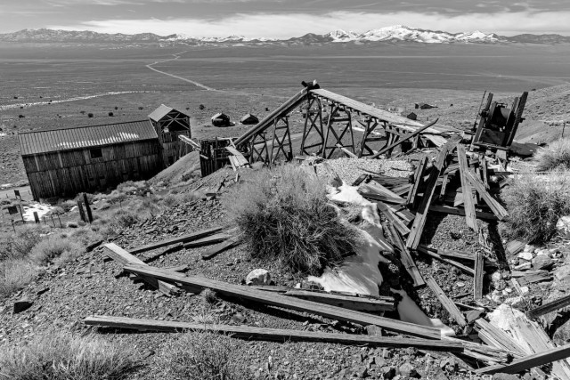 A black and white landscape photo of ruins at a ghost town in central Nevada. From the bottom of the frame to the top (front to back) we see a scattered pile of weathered lumber in the foreground. It sits on a hill with a couple of sagebrush plants. A trestle for mining carts to delivery ore to the stamp mill runs from center-right and down the hill to center frame and enters a large wood barn like building. In the distance, the middle ground, is a very large flat valley with a dirt road winding off toward distant snow covered mountains in the background. A thin strip of sky is seen above the mountains. There's a cloud bank just above the mountains.