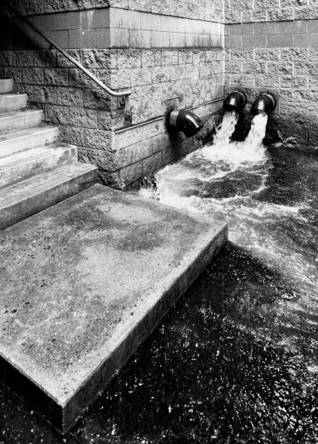Steps descend from an indoor parking structure to a concrete landing. Rainwater pours out of two nearby downspouts flooding the street around the concrete landing.