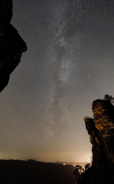 Hochkant-Fotografie der Milchstraße. Das Bild ist eingerahmt von Felsen in der oberen linken und unteren rechten Ecke, wobei vor allem der Rechte von unten sanft gelblich angestrahlt wird. Im Zentrum steht das Milchstraßenband, welches sich nahezu senkrecht aus dem Horizont am unteren Bildrand erhebt und über das ganze streckt. Die Strukturen sind deutlich zu erkennen und auch die Farben sind zu erahnen. Am Horizont unten steht ein schmales Wolkenband zwischen gelber Lichtverschmutzung. Man kann ein Tal erahnen, in dem einige Lichter leuchten.