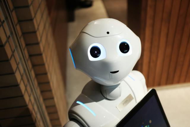 A white humanoid robot with large, glowing blue eyes looks up toward the camera. Its smooth plastic face has a small, friendly smile. Soft blue accent lights run along its head and shoulders. It holds a tablet in front of its chest, and the background shows a tiled wall and warm wooden paneling.
