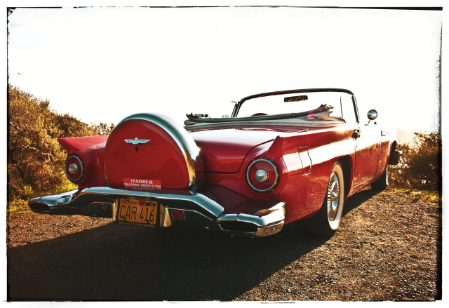 A very red mid-1950s Ford Thunderbird convertible, with stylish chrome trim, smallish rear fins, and a giant spare tire compartment above the rear bumper. Photographed from the rear at a 45° angle behind the right tail light, the car is stunning. It stands there in full glory, shining in the sun, top down, on a dirt patch of ground facing the Pacific Ocean. The white wall tires are a nice, and necessary, touch. 

Note: there's a bumper sticker on the rear tire compartment that says, in large white letters, I'D RATHER BE SMASHING IMPERIALISM.

We all would be, car. We all would be.