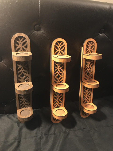 3 handmade laser-cut wood wall sconces in L-to-R Dark Walnut, Cherry, and Cherry/Walnut two tone. They are tall and narrow with rounded tops, bottoms and candle shelves, and feature a layered geometric floral pattern behind the candles.