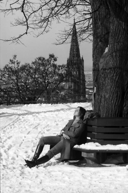 Schwarz-weiß Aufnahme: Freiburg 18. Februar 1969. Sonnendes Mädchen am Kanonenplatz im Schnee auf einer Bank sitzend, zurückgelehnt, die Augen geschlossen. Im Hintergrund der Turm des Münsters