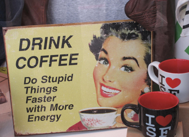 A  plaque, showing some wear, which says:
Drink coffee.
Do stupid things faster with more energy.
To the right of the words is a picture of a smiling woman holding a cup of coffee.

Beside the plaque are a couple I ❤️ SF mugs. 
