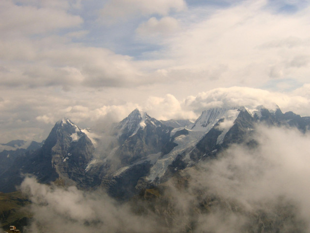 A view of three mountains, the Eiger, Mönch and Jungfrau, ascending in height from left to right. Each has dark shadows on the north (left) side and are capped with snow. The rock of the mountains appears dark blue. In front there is quite thick low cloud obscuring the lower levels. Above are both fluffy and wispy white clouds and smatterings of blue sky.