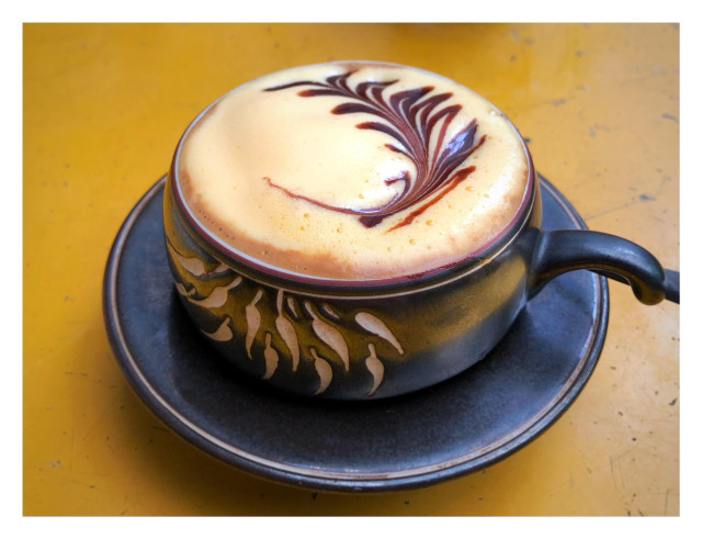 Photo of a cup of coffee on a blue saucer atop a saffron-colored table, viewed from slightly above. The cup is made of dark brown clay or ceramic with decorative light-colored leaf silhouettes on the side. The coffee is topped with a light, frothy cream. A dark brown floral, leafy design has been “drawn” into the foam by the barista.