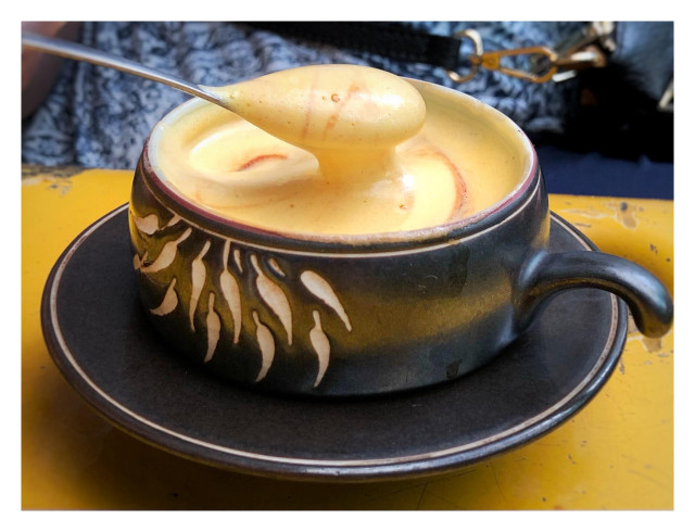 Another view of the same cup of coffee as in the previous image. The latte art has been stirred into the drink, and a spoon is lifting some of the cream out, demonstrating how thick it is. In the background is a blurred glimpse of a patterned blue shirt and a black handbag.