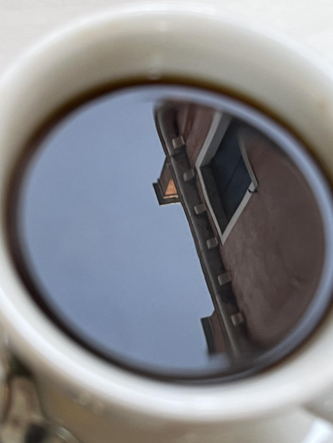 Closeup photo looking straight down into my morning cup of coffee, sitting outside my local bar. The rim of the white china cup is slightly out of focus, and on the right half of the glossy dark still surface of the coffee within there is the sharp reflection of the top of a house: red-brown wall, a square window with closed dark wooden shutters, and two chimney stacks, one of which is luminous in the light of the morning sun. 