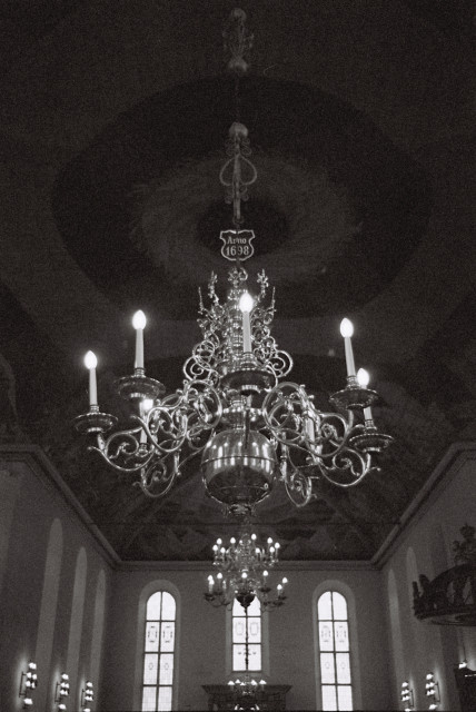 A grainy, black-and-white photo of a chandelier in Oslo Cathedral. The chandelier is not enormous, but it is very elaborate, with curly, organic-looking arms and decorations shining in the light from its now electric candles. At the top hangs a small shield saying ‘Anno 1698’. In the background is the cathedral's painted ceiling, some stained-glass windows and another chandelier on the other side of the nave.