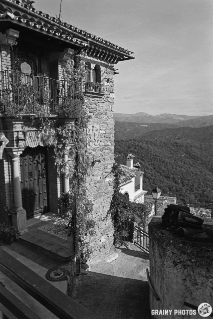 An old stone building with elaborate architecture and metal balconies overlooks a mountainous landscape. The scene features lush greenery and a distant view of rolling hills under a clear sky, creating a serene and picturesque ambiance.