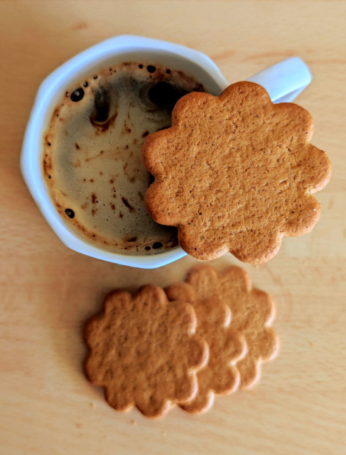 Photo taken from above of an octagonal mug with slightly frothy black coffee within and a thin ginger biscuit sort of in the shape of a chubby nine-pointed star on top. There are three more biscuits overlapping below, on a pale wooden surface.