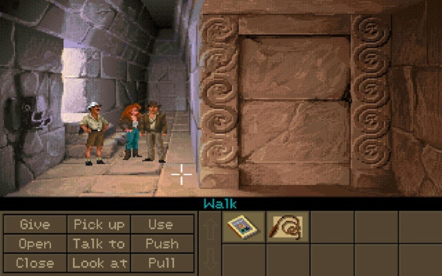 In-game screenshot of Indiana Jones and the Fate of Atlantis. It shows the inside of a temple with beige stone walls and stone floor in a pixelized art style. Three people are standing in the room (Indiana Jones in brown leather clothing, a woman with long red hair wearing blue trousers and another man wearing a tropical explorer outfit).
The bottom quarter of the screen is filled with activity verbs like give, open, push or pull as well as items the player has collected. A magazine and Indy's legendary whip in this case.
