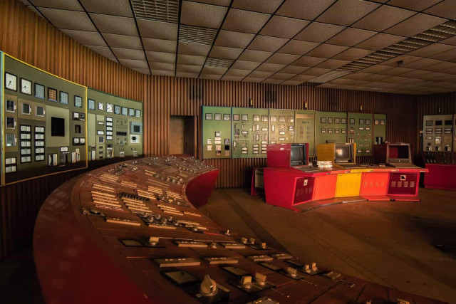 Large, dimly lit control room with curved consoles and multiple analog dials, switches, and monitors along wood-paneled and green walls.