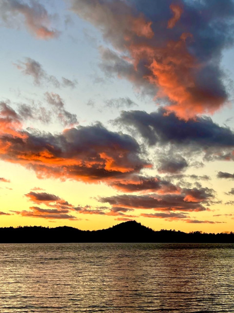 Large individual clouds, colored deep red by the sunset, hover over a lake like invading UFOs. The sky is blue near the top, and yellow near the horizon. The silhouettes of sand dunes are on the far side of the lake.