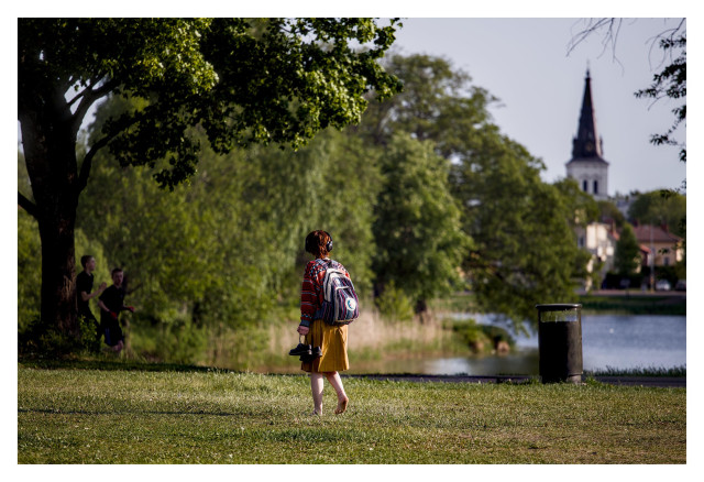 En liggande bild på en tjej med ryggsäck och gul kjol och skorna i handen som promenerar över en gräsmatta och tittar över ett tjärn bortåt kyrkan på andra sidan. Två killar kommer joggande i vänster bildkant.