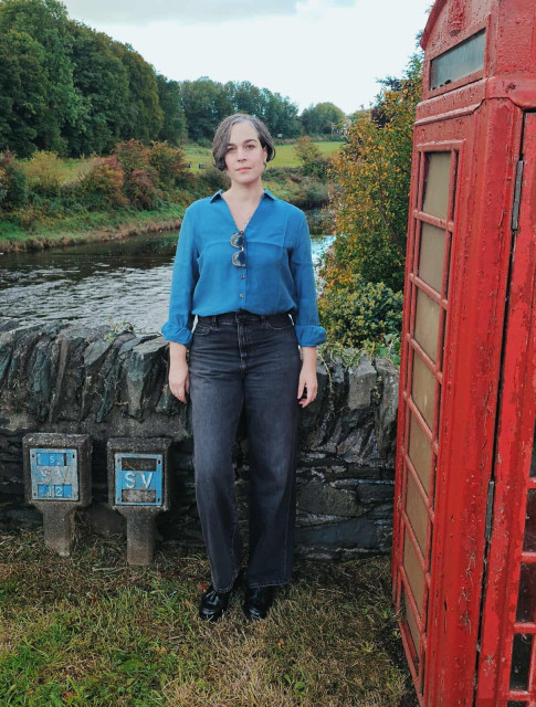 A woman with short, greying dark hair stands outdoors beside an old red British phone box. She wears a teal button-up shirt tucked into high waisted dark jeans and black shoes. A pair of sunglasses hangs from her shirt. Behind her is a stone wall, a flowing river, and trees with early autumn foliage. Two small blue andwhite utility markers are set into the grass near her feet. The atmosphere is calm and slightly overcast.