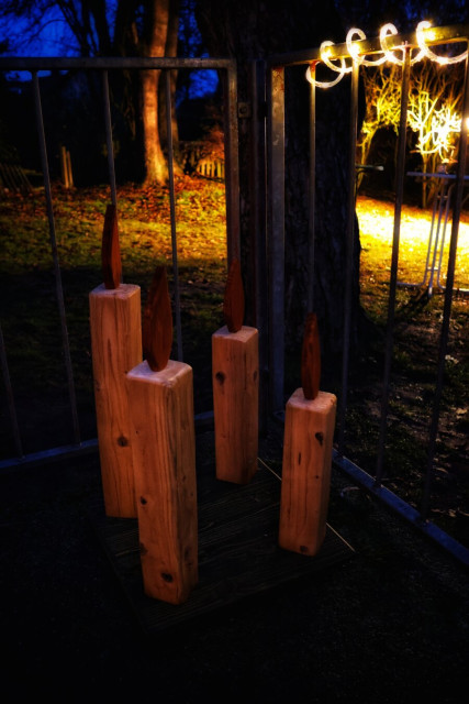A wooden Advent wreath featuring four rustic wooden candles. The wreath is placed outdoors on a dark surface, with a metal railing in the background. The scene is set in the evening, with decorative lights wrapped around the railing, creating a warm and festive atmosphere.