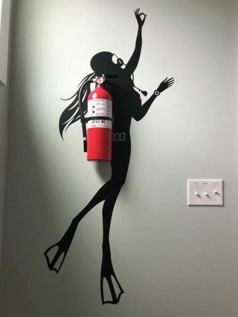 A female diver/frogman is painted on the wall as a black outline in such a way that the wall mounted fire extinguisher appears to be her oxygen tank