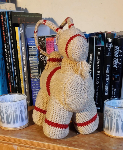 A crocheted Yule goat, with red bands on its legs, belly, nose and tail. It's standing on a bookshelf with tall books behind it. 