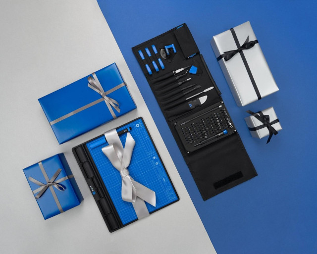An iFixit repair mat and fully opened tool roll are arranged on a split blue-and-gray background alongside neatly wrapped silver and blue gifts with black and silver ribbons.