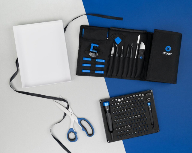 An open iFixit tool roll and a full bit driver kit are arranged on a split blue-and-gray background next to gift-wrapping supplies, including scissors, ribbon, and an empty gift box.