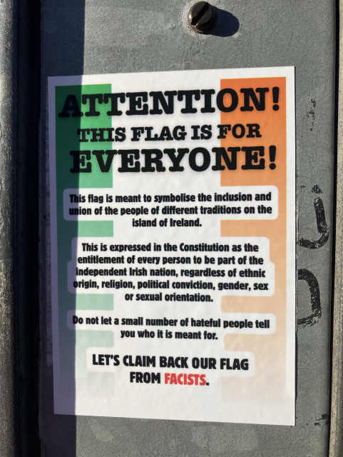 Photo of a sticker on a pedestrian crossing pole in Cabra, Dublin. It has a background with the colours of the tricolour flag of Ireland: green, white, and orange. The text reads: 

"ATTENTION!

THIS FLAG IS FOR EVERYONE!

This flag is meant to symbolise the inclusion and union of the people of different traditions on the island of Ireland.

This is expressed in the Constitution as the entitlement of every person to be part of the independent Irish nation, regardless of ethnic origin, religion, political conviction, gender, sex or sexual orientation.

Do not let a small number of hateful people tell you who it is meant for.

LET'S CLAIM BACK OUR FLAG
FROM FASCISTS."